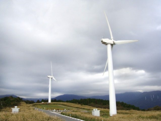 曇り空のなか、勢いよく回転する伊豆町の町営風車。右奥の山肌に小さく見える白い柱は、熱川の風車。写真:寺倉聡子
