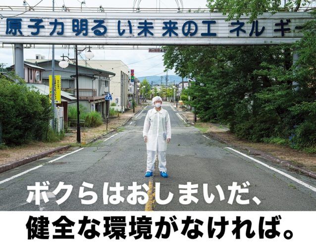 ひとけのない双葉町で、スローガンが掲げられた看板の前に立つ、防護服とマスクを身に着けた人。2011年3月、福島第一原子力発電所の事故により全町民が避難した。このスローガンは今、皮肉にも原子力への依存の危険性を思い出させている。Photo: Arkadiusz Podniesinski
